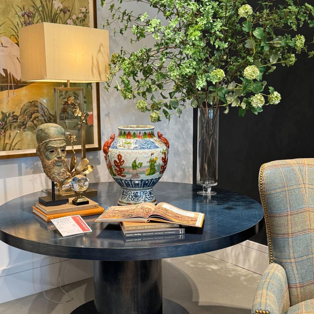 Navy pedestal table styled with a famille-rose vase, sculpture, books and greenery beside a plaid armchair